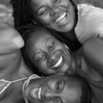 A black-and-white photo of three friends smiling and laughing, laying closely together outdoors with relaxed, joyful expressions. Two are wearing sleeveless tops, while the third has braided hair and a visible hoop earring. All appear happy and carefree, capturing the spirit of Playa del Carmen.