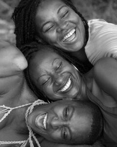 A black-and-white photo of three friends smiling and laughing, laying closely together outdoors with relaxed, joyful expressions. Two are wearing sleeveless tops, while the third has braided hair and a visible hoop earring. All appear happy and carefree, capturing the spirit of Playa del Carmen.