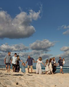 A multi-generational family portrait unfolds as a group of people, including adults and children, hold hands while strolling along a sandy beach. The sky is partly cloudy, and the ocean waves can be seen in the background, hinting at their shared enjoyment of a sunny day together.